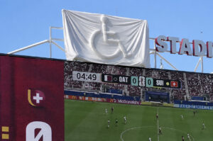 Gran lona blanca cubre un logotipo del símbolo internacional de accesibilidad sobre una pantalla de un estadio mientras se disputa un partido de fútbol. La imagen ilustra el paralelismo del artículo: intentar ocultar algo puede acabar haciéndolo más visible, como ocurre con las barreras de accesibilidad.