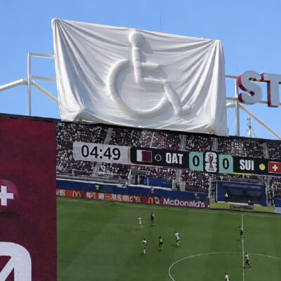 Gran lona blanca cubre un logotipo del símbolo internacional de accesibilidad sobre una pantalla de un estadio mientras se disputa un partido de fútbol. La imagen ilustra el paralelismo del artículo: intentar ocultar algo puede acabar haciéndolo más visible, como ocurre con las barreras de accesibilidad.