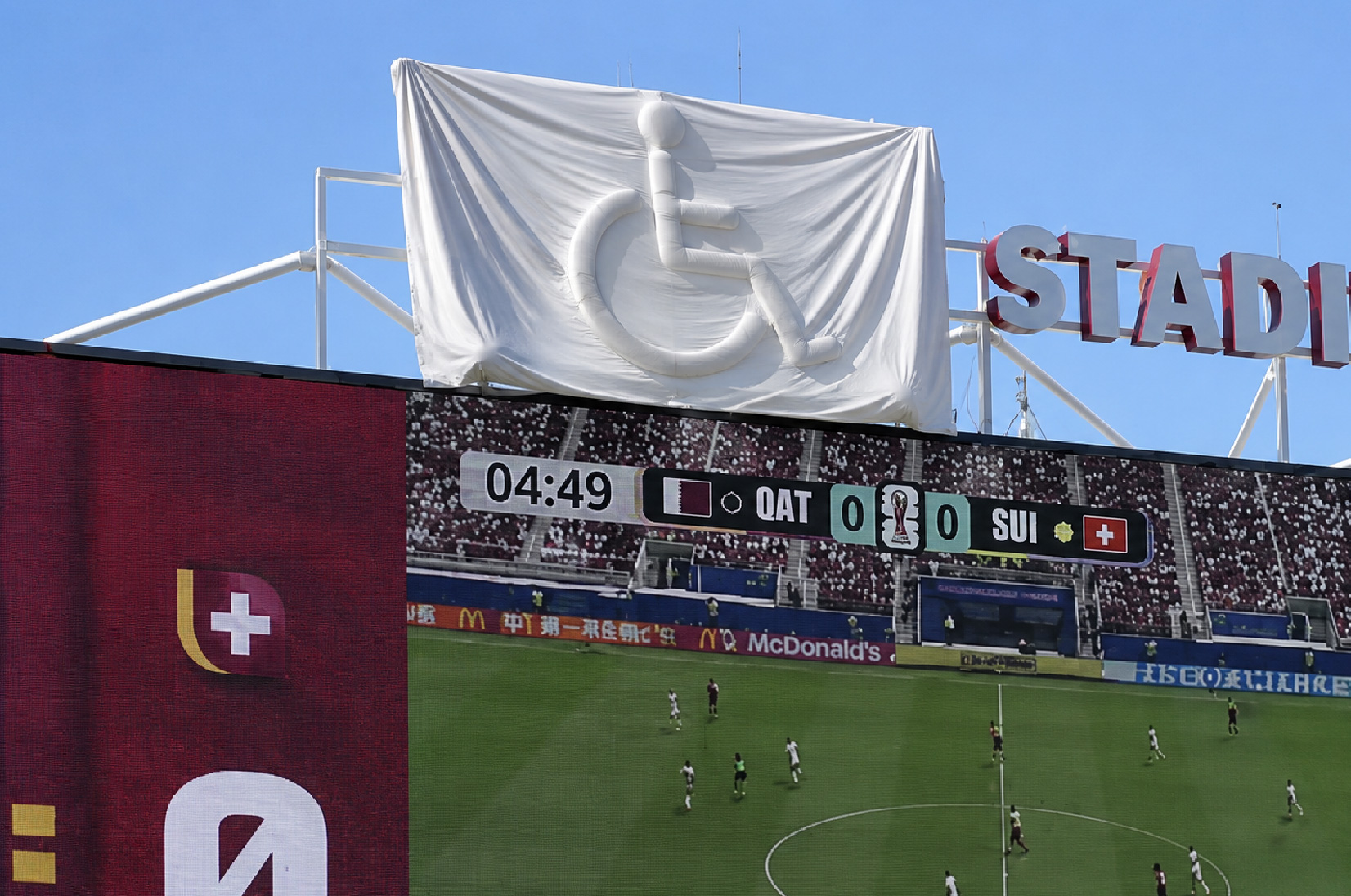 Gran lona blanca cubre un logotipo del símbolo internacional de accesibilidad sobre una pantalla de un estadio mientras se disputa un partido de fútbol. La imagen ilustra el paralelismo del artículo: intentar ocultar algo puede acabar haciéndolo más visible, como ocurre con las barreras de accesibilidad.