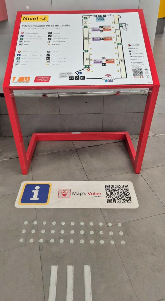 Nuevo plano tacto-visual accesible del Intercambiador de Plaza de Castilla, instalado sobre un atril rojo y conectado mediante pavimento táctil y botones de señalización. Frente al plano, colocado en el suelo, hay un vinilo adhesivo con el pictograma de información y un código QR de Map’s Voice de Puntodis.