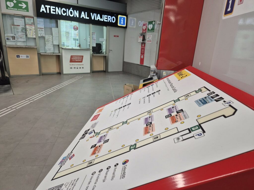 Plano tacto-visual instalado sobre un atril rojo en la planta -2 del Intercambiador de Plaza de Castilla, situado frente al punto de Atención al Viajero del Consorcio Regional de Transportes de Madrid. En el suelo se aprecia un encaminamiento que conduce hasta el plano. El recurso de señalización accesible incorpora relieve, braille, alto contraste, pictogramas y un código QR con acceso a Map's Voice, facilitando la orientación de las personas usuarias.