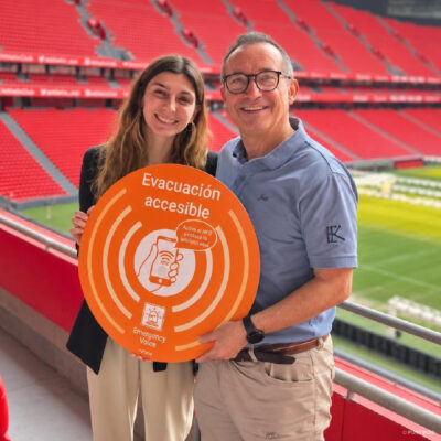 Andrea Barranco, a la izquierda, y Jon Ibarra, a la derecha, posan sosteniendo un vinilo circular naranja de Emergency Voice, con letras blancas, ondas concéntricas e indicaciones para consultar la evacuación accesible mediante NFC. Al fondo se ven las gradas rojas y el césped de San Mamés.