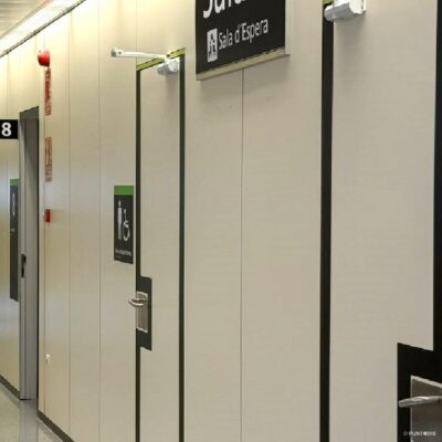 Pasillo del área de Oftalmología del Hospital Universitario en Lleida, con bandas negras de encaminamiento tacto-visual en el suelo y banderolas con números blancos que identifican las consultas. A la derecha, puertas delimitadas con franjas negras y señales de alto contraste con pictogramas, texto y braille para identificar los aseos adaptados.