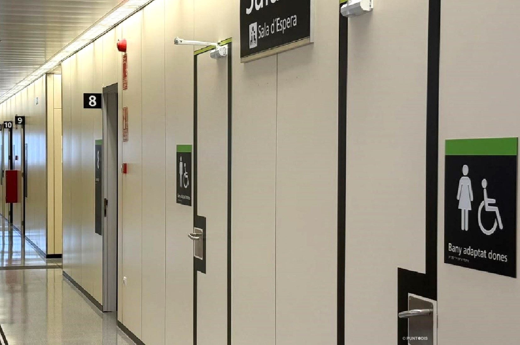 Pasillo del área de Oftalmología del Hospital Universitario en Lleida, con bandas negras de encaminamiento tacto-visual en el suelo y banderolas con números blancos que identifican las consultas. A la derecha, puertas delimitadas con franjas negras y señales de alto contraste con pictogramas, texto y braille para identificar los aseos adaptados.